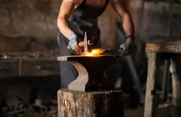 Blacksmith forging metal on an anvil with sparks flying in a blacksmith workshop.