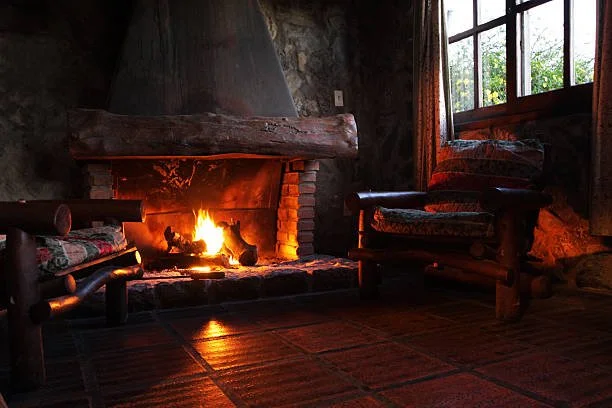 Cozy living room with a lit fireplace, wooden furniture with cushioned seats, and a large window showing greenery outside.