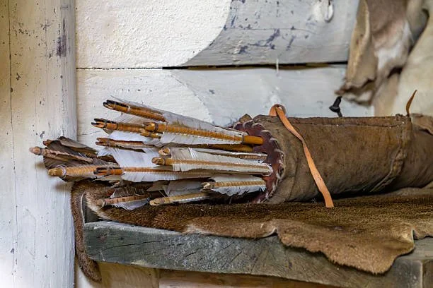 A bundle of traditional Native American arrows with feather fletching, resting on a leather quiver and a fur-lined surface inside a rustic wooden structure.