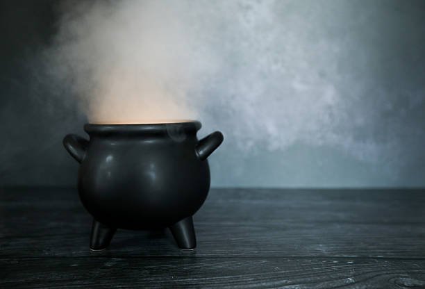 A black cauldron with steam rising from it on a dark wooden surface against a gray textured background.