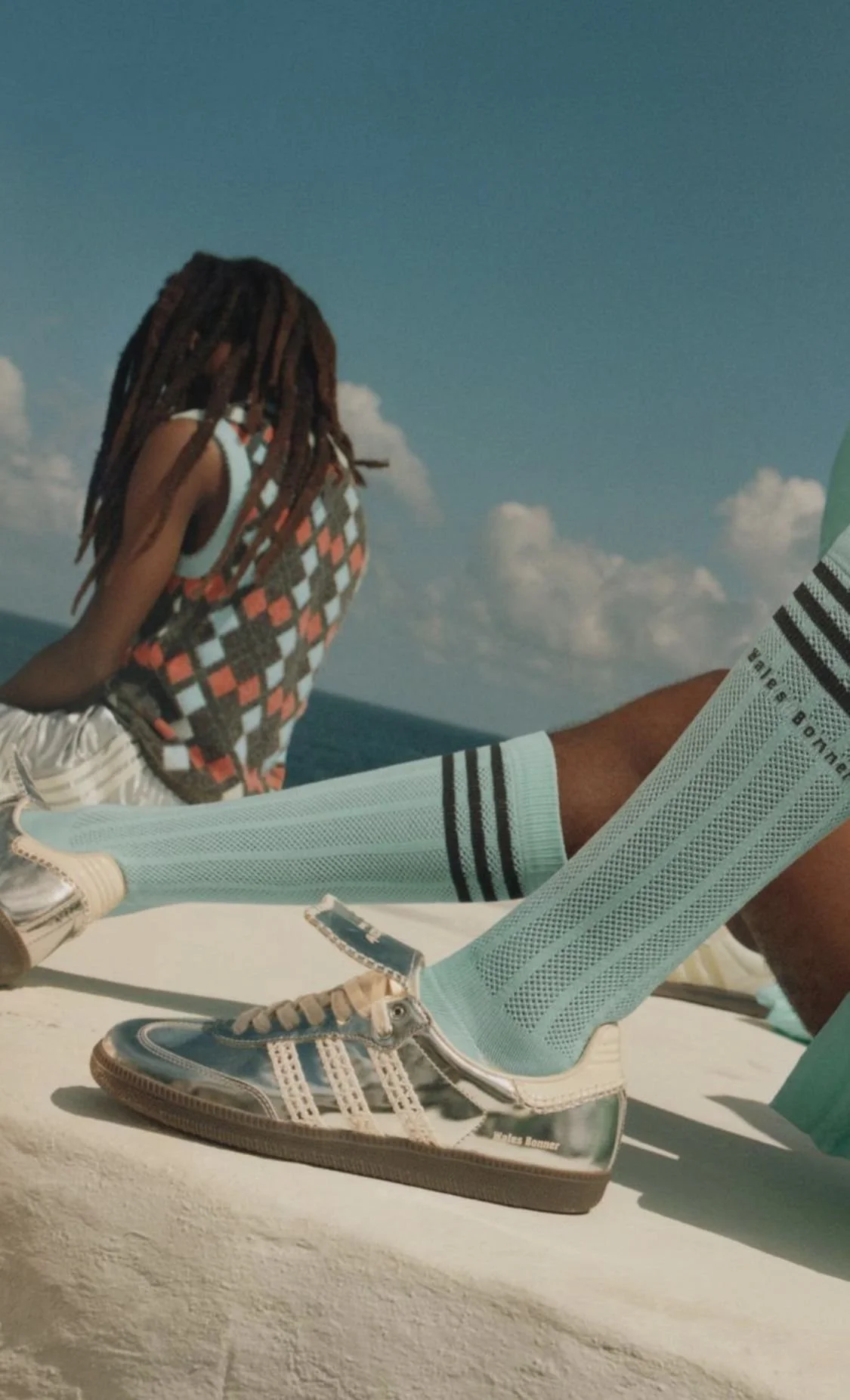 A person wearing shiny silver Adidas sneakers, light blue socks with black stripes, and beige shorts, sitting on a white surface near the ocean with a person in the background, sky with clouds.
