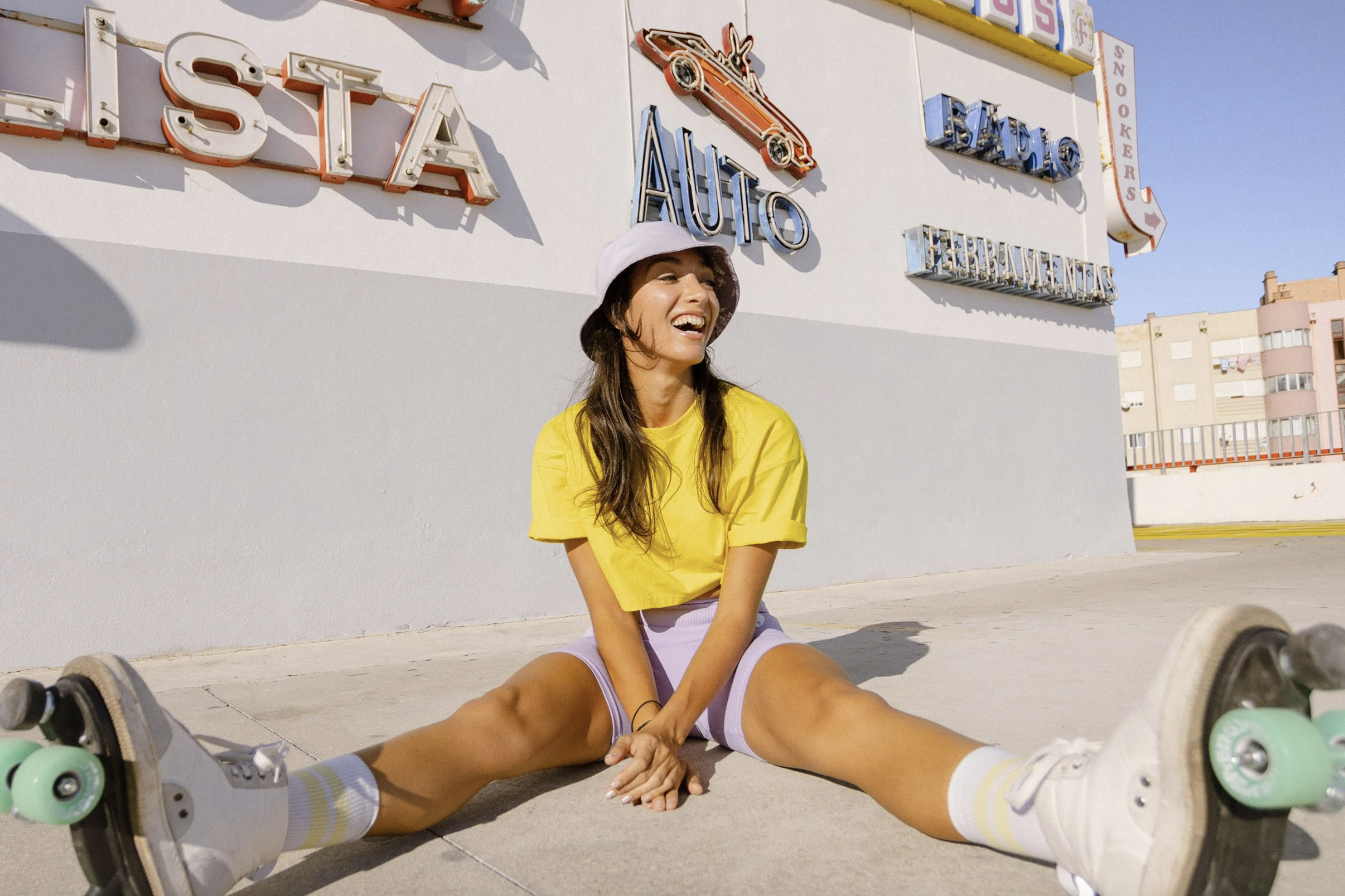 A young woman sitting on the pavement in front of a building with multiple vintage neon signsShe is wearing a yellow T-shirt, light purple shorts, white sneakers, striped socks, and a light pink bucket hat. She appears to be happy and relaxed, with a bright smile.