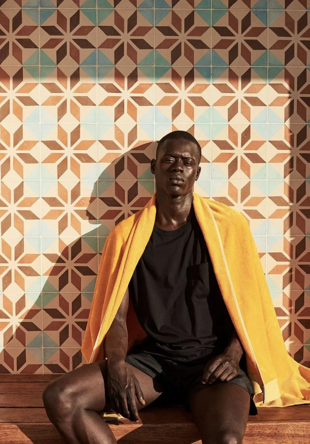A young man sitting on wooden steps against a colorful geometric tile wall, wearing a black shirt and shorts with a yellow towel draped over his shoulders, looking directly at the camera.