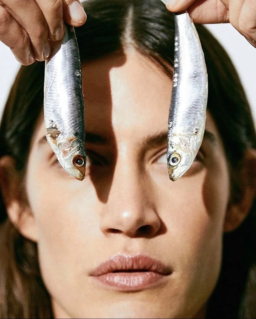 A fashionable woman in Lisbon with dark hair holding two small fish in front of their face, covering their eyes, with a shadow cast across the person's face.