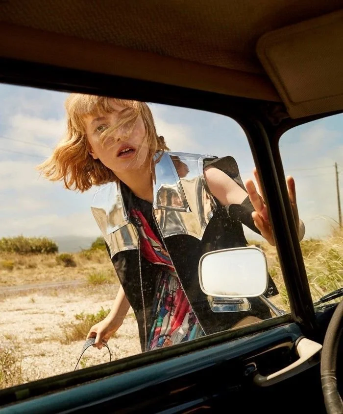Young girl with blonde hair looking into a vehicle through the window on a sunny day, outdoors in a desert-like area.
