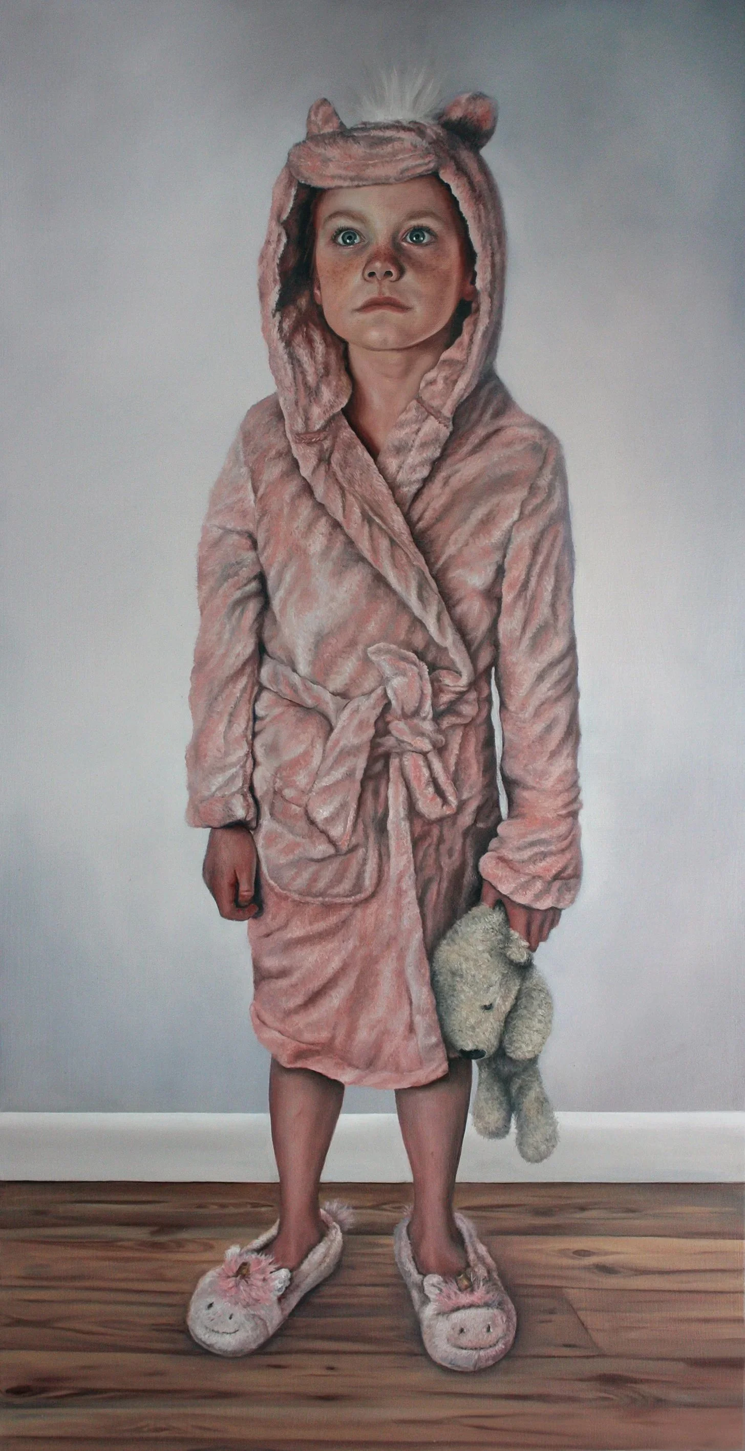 A young girl wearing a pink fluffy bathrobe with a hood shaped like a bear's head, holding a teddy bear, and wearing matching bear slippers. She stands on a wooden floor against a plain wall, looking slightly upward.
