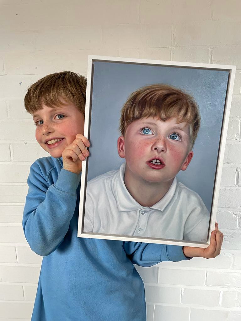 A boy holding a framed portrait of himself, which depicts him with bright blue eyes, freckles, and a surprised expression.