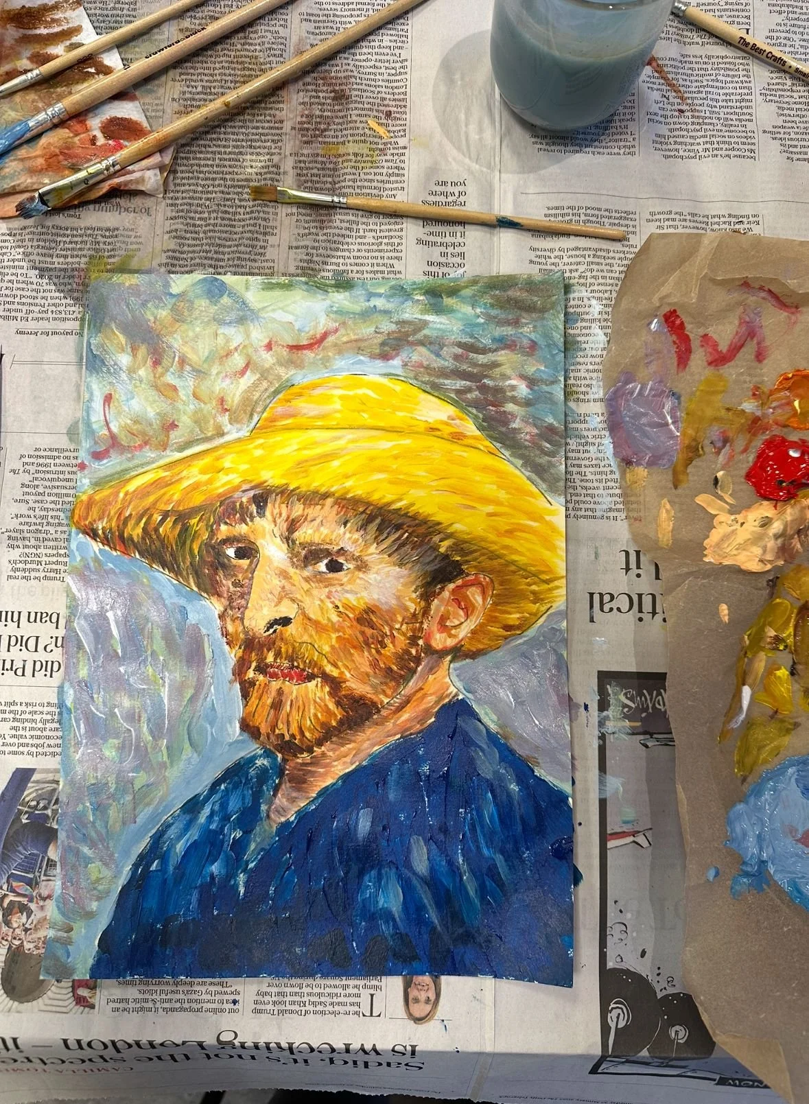 Oil painting of a man with a beard wearing a large yellow hat and a blue shirt, set on a table covered with newspapers, paintbrushes, and a palette with paint.