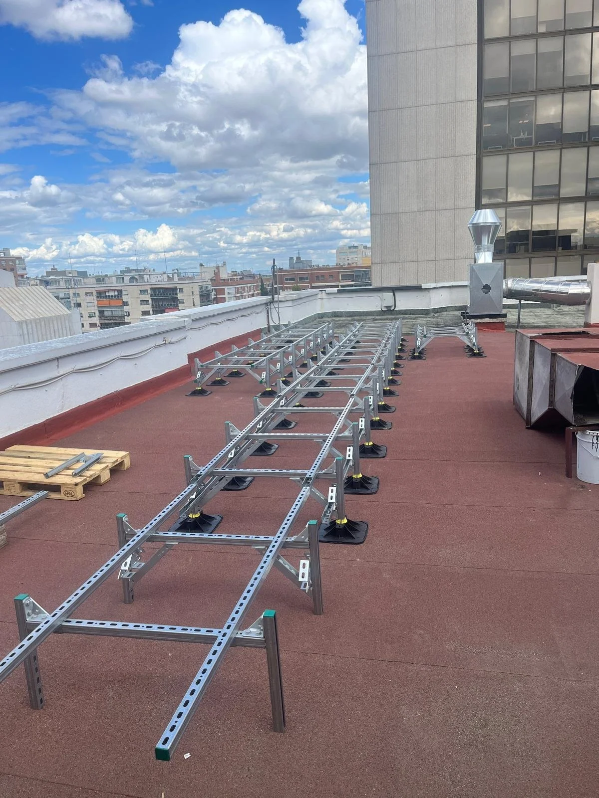 Estructuras de metal instaladas en un techo plano, junto a una máquina de ventilación, con edificios urbanos y cielo con nubes en el fondo.
