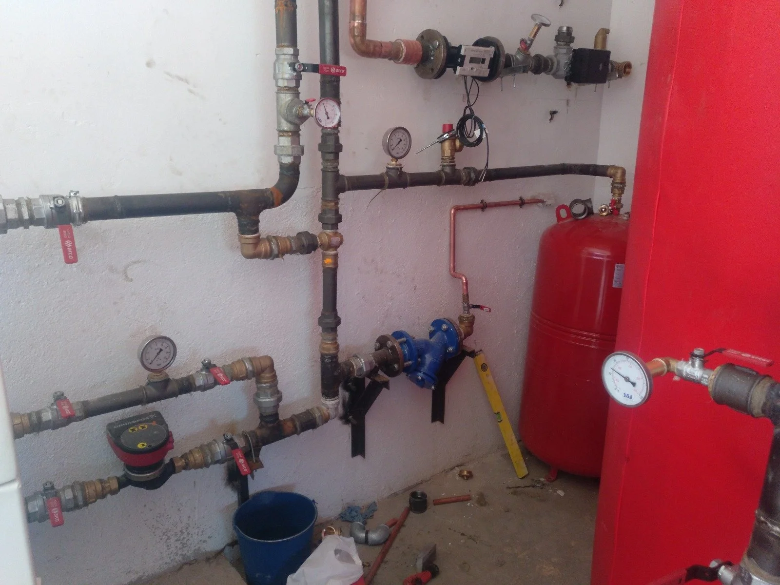 Sistema de instalaciones hidráulicas con tuberías, válvulas, medidores y un tanque de expansión rojo en una pared en construcción.