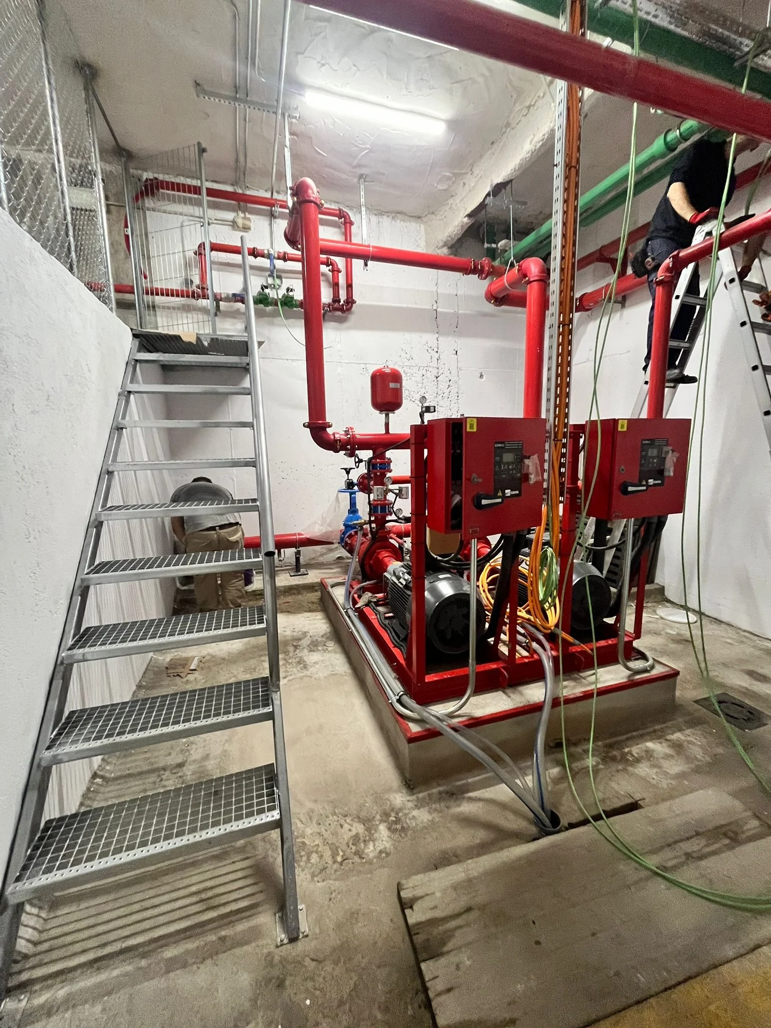 Sistema de bombeo contra incendios con tuberías rojas, bomba, válvulas, paneles de control y escaleras en un espacio subterráneo.