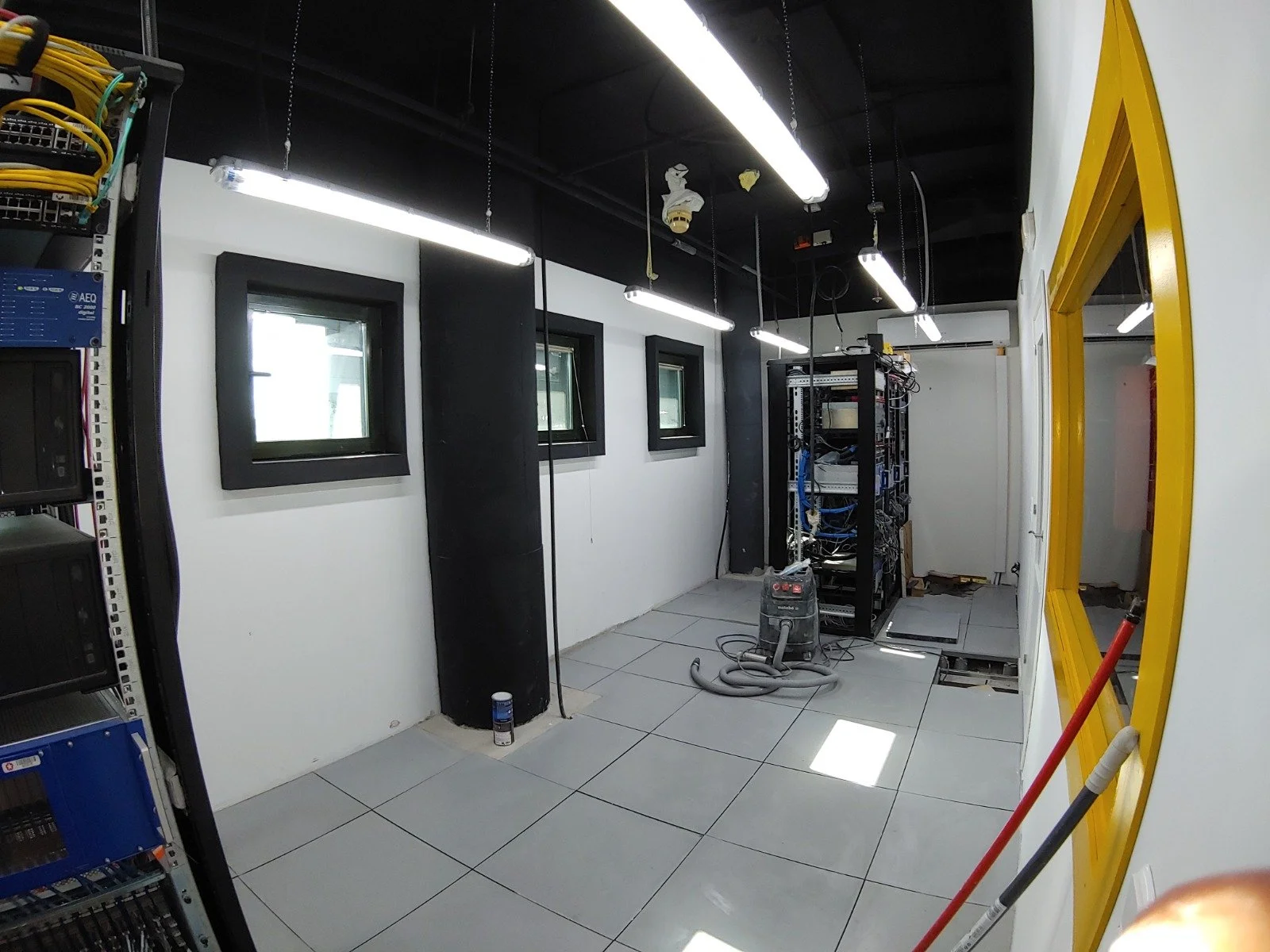 Sala de servidores en construcción o mantenimiento con equipo de cableado, una estantería con componentes y ventanas pequeñas.