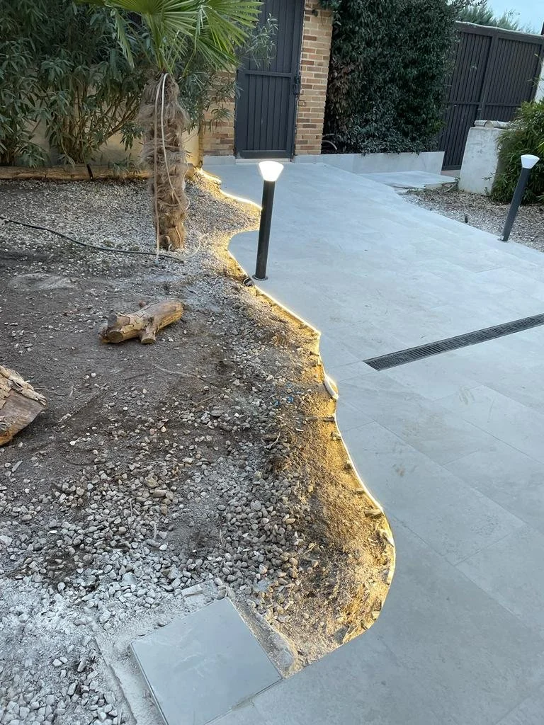 Entrada de un jardín o patio con piso de adoquines y área de tierra con plantas, con iluminación decorativa en el borde. Entrada con puerta de madera y cerca de muro de ladrillo.
