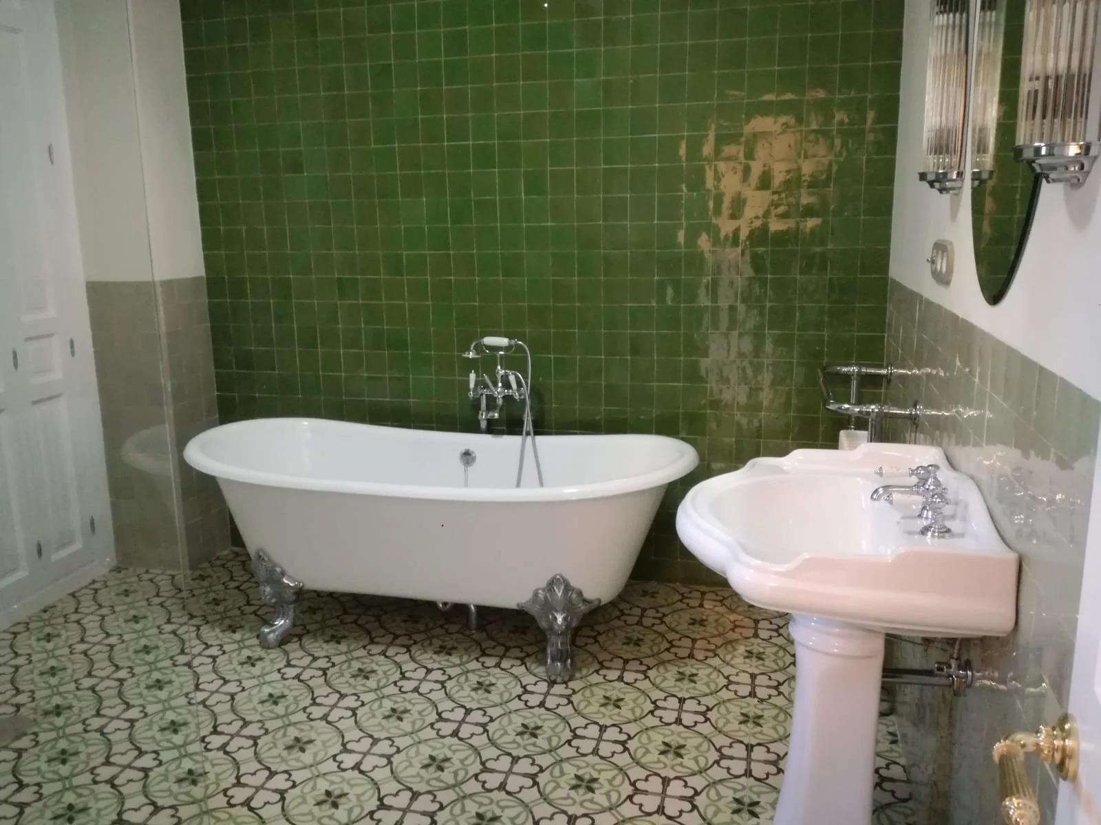 Baño con pared de azulejos verdes, tina de patas de diseño clásico, lavabo con pedestal y espejo circular.