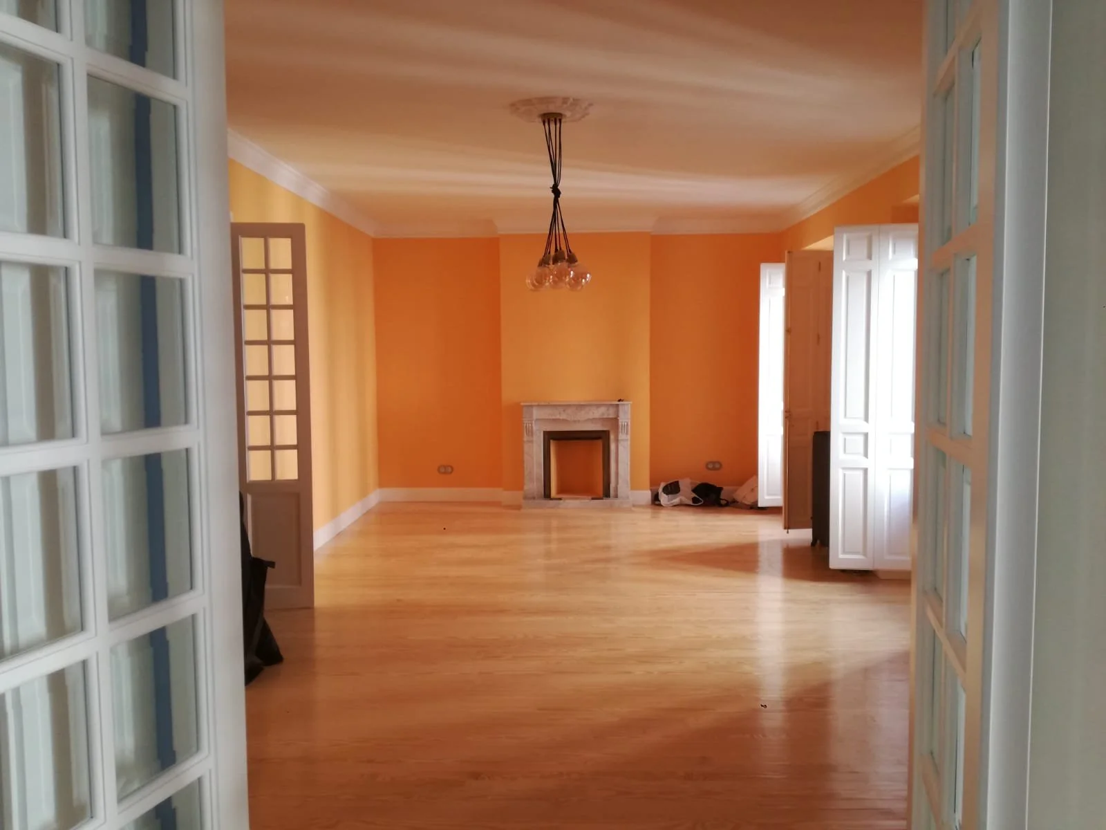 Vista del interior de una sala vacía con paredes naranjas, piso de madera clara, chimenea de piedra en el centro y ventanas blancas abiertas, vista desde una puerta de vidrio.