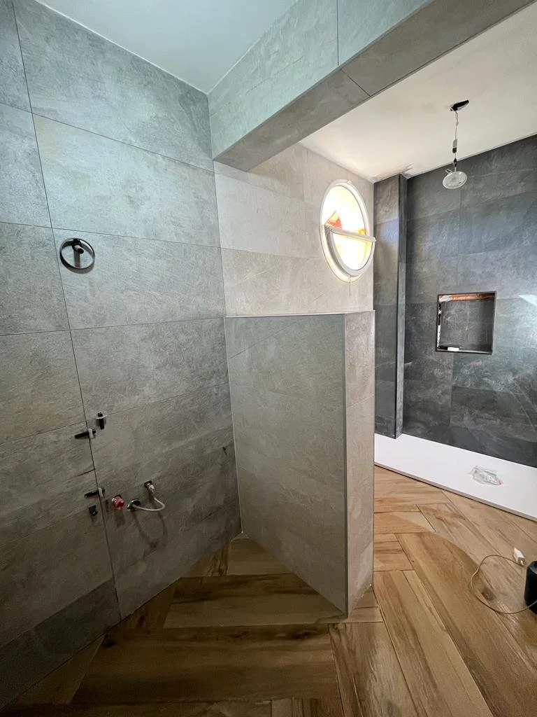 Sala de baño en construcción con paredes de azulejo gris, una ventana redonda y suelo de madera, sin accesorios ni mobiliario instalado.