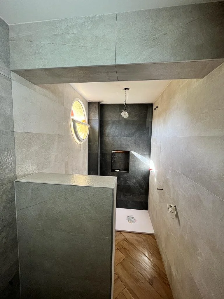 Baño en construcción con paredes de azulejo gris y suelo de madera, con una ventana redonda y accesorios en proceso de instalación.