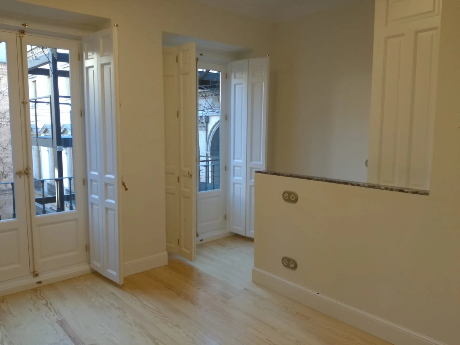 Vista de una habitación vacía con pisos de madera clara, paredes blancas y grandes ventanas con puertas de madera blancas que dan a un balcón con barandales negros.