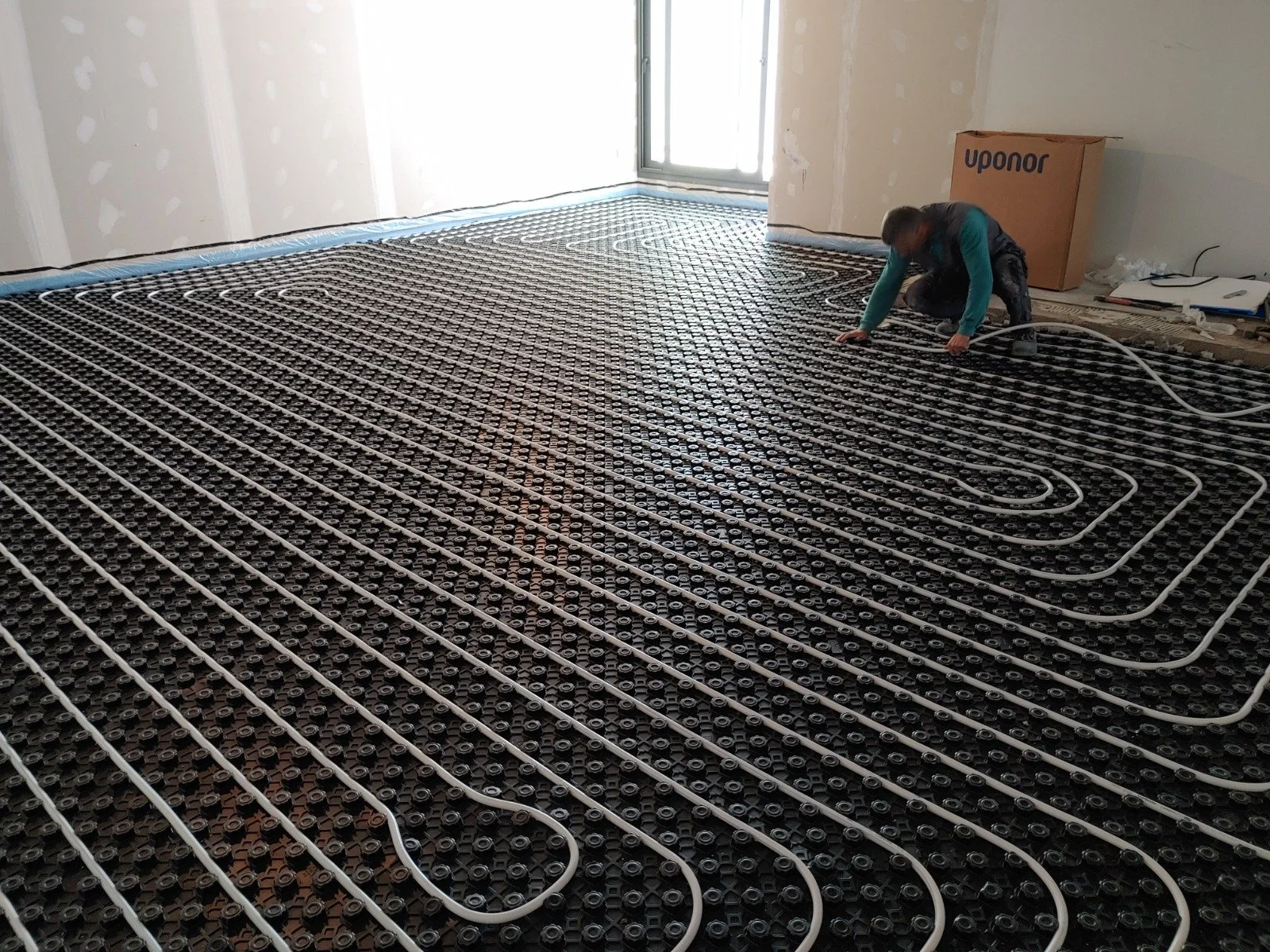 Un trabajador instalando sistema de calefacción por suelo radiante en una habitación en construcción con paredes de drywall y una ventana.