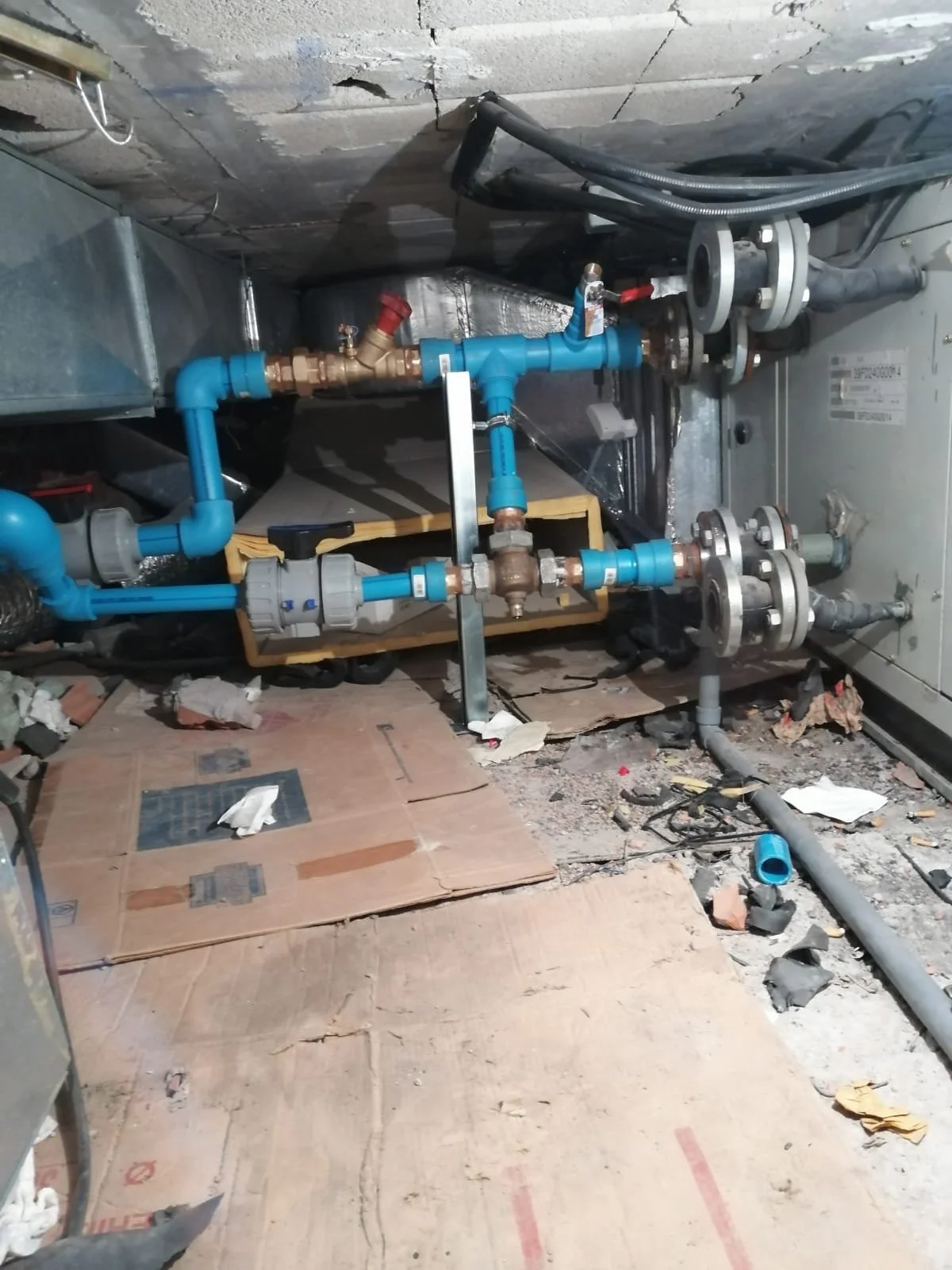 Sistema de tuberías y válvulas en un área de mantenimiento, con tuberías azules, conexiones metálicas, y cables en el techo, sobre un suelo de cartón y escombros.