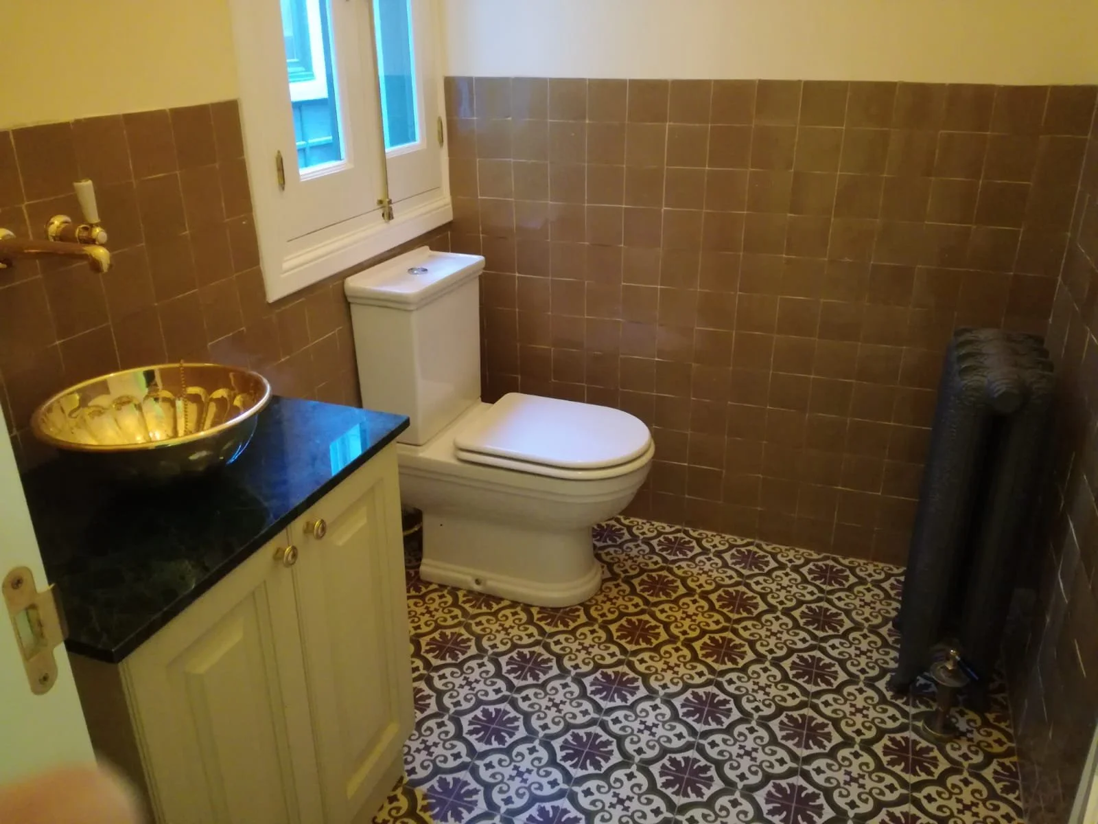 Baño con azulejos en la pared, inodoro, lavabo con lavabo de oro y encimera negra, radiador negro, ventana y espejo.