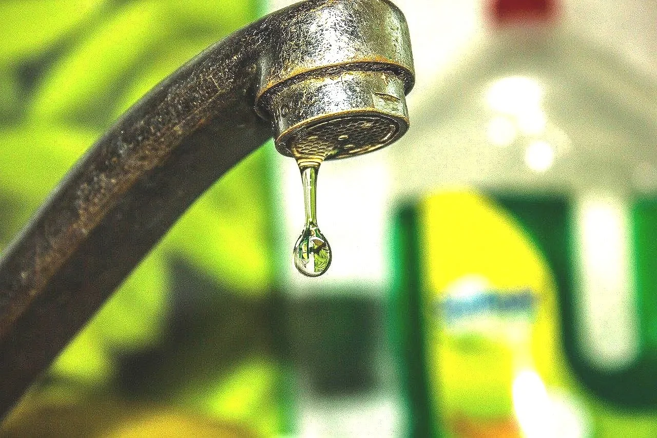 Close-up of a rusty faucet with a single droplet of water hanging from it, blurred background with green and yellow colors.