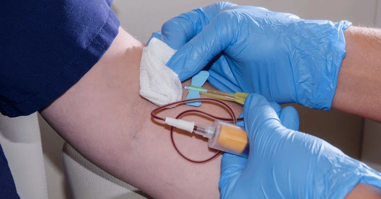 Medical professional wearing blue gloves drawing blood from a patient's arm with a syringe connected to a blood collection tube.