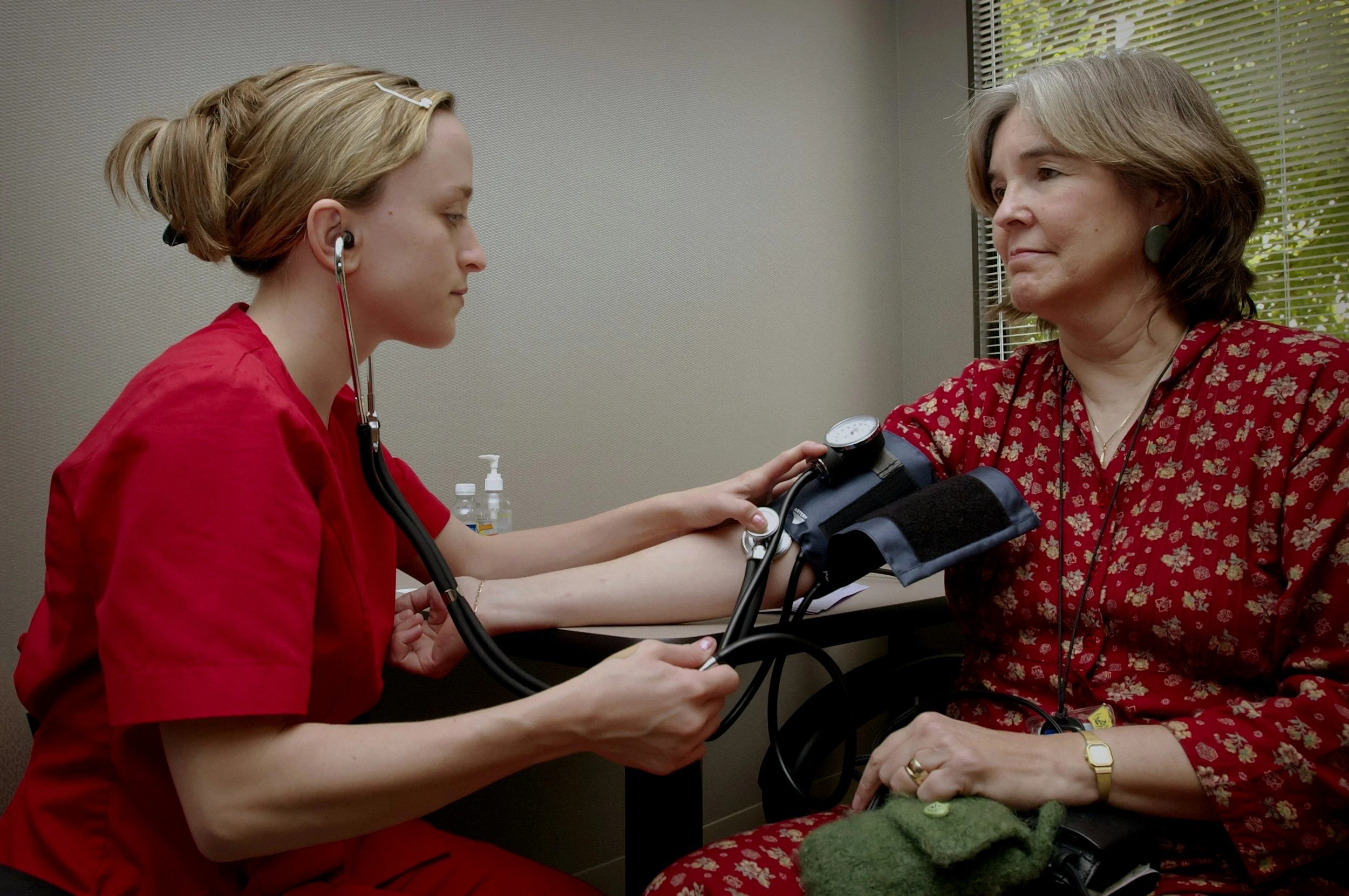 A healthcare professional, wearing red scrubs, is using a blood pressure monitor on a middle-aged woman with gray hair and a red floral shirt during a medical check-up.
