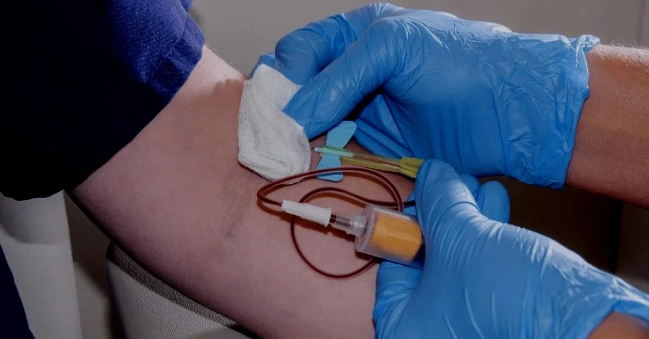 Medical professional in blue gloves draws blood from a patient's arm using a needle and tubing.