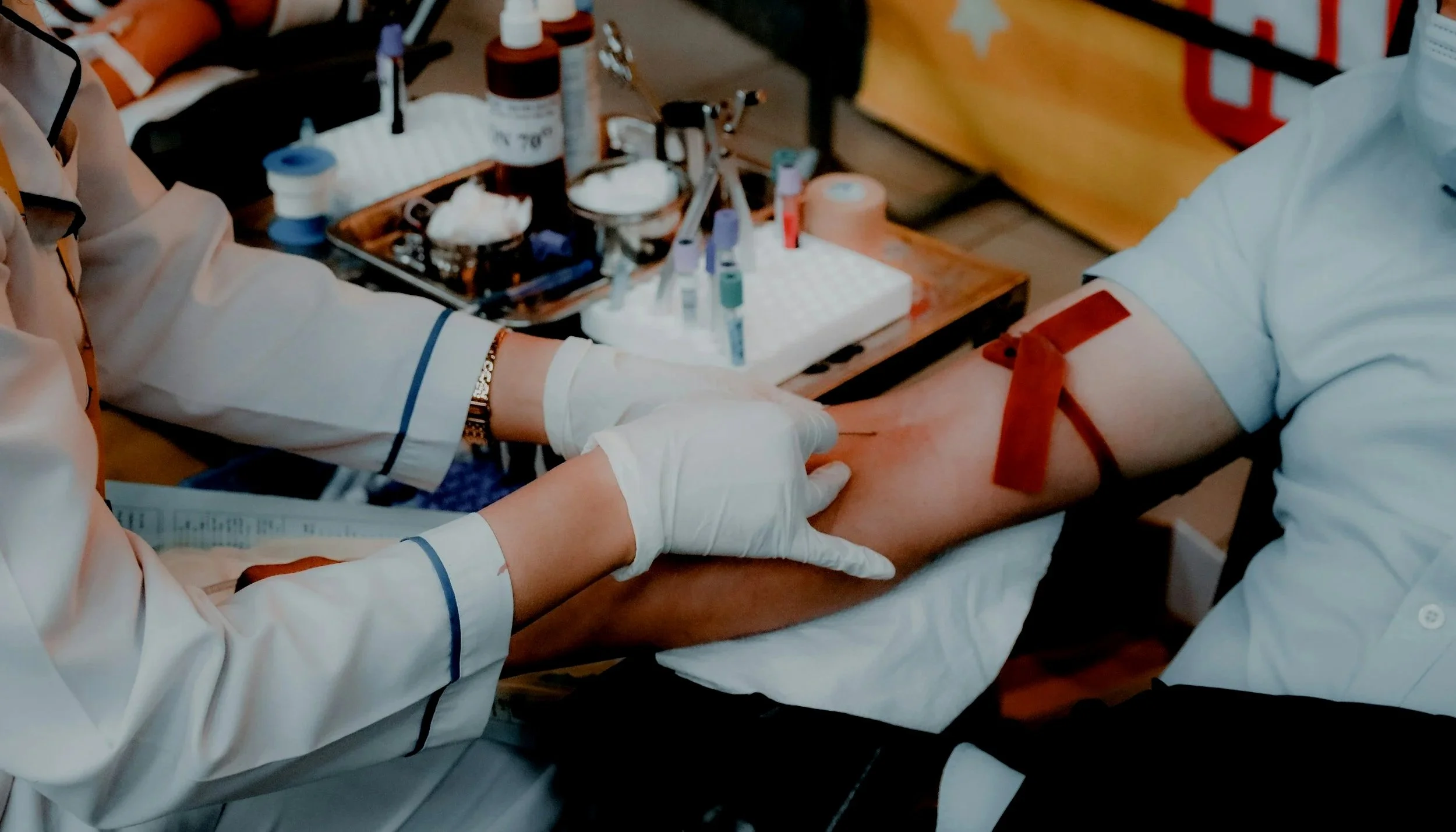 A person is donating blood, with a healthcare worker preparing to draw blood from their arm. The healthcare worker is wearing gloves, and the patient's arm has a bandage around the puncture site. Medical supplies and equipment are visible on a table in the background.
