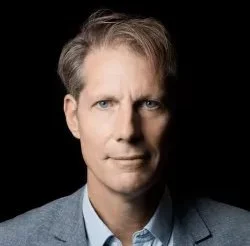 Portrait of a man with light brown hair, blue eyes, and fair skin, wearing a light blue shirt and gray blazer, against a black background.