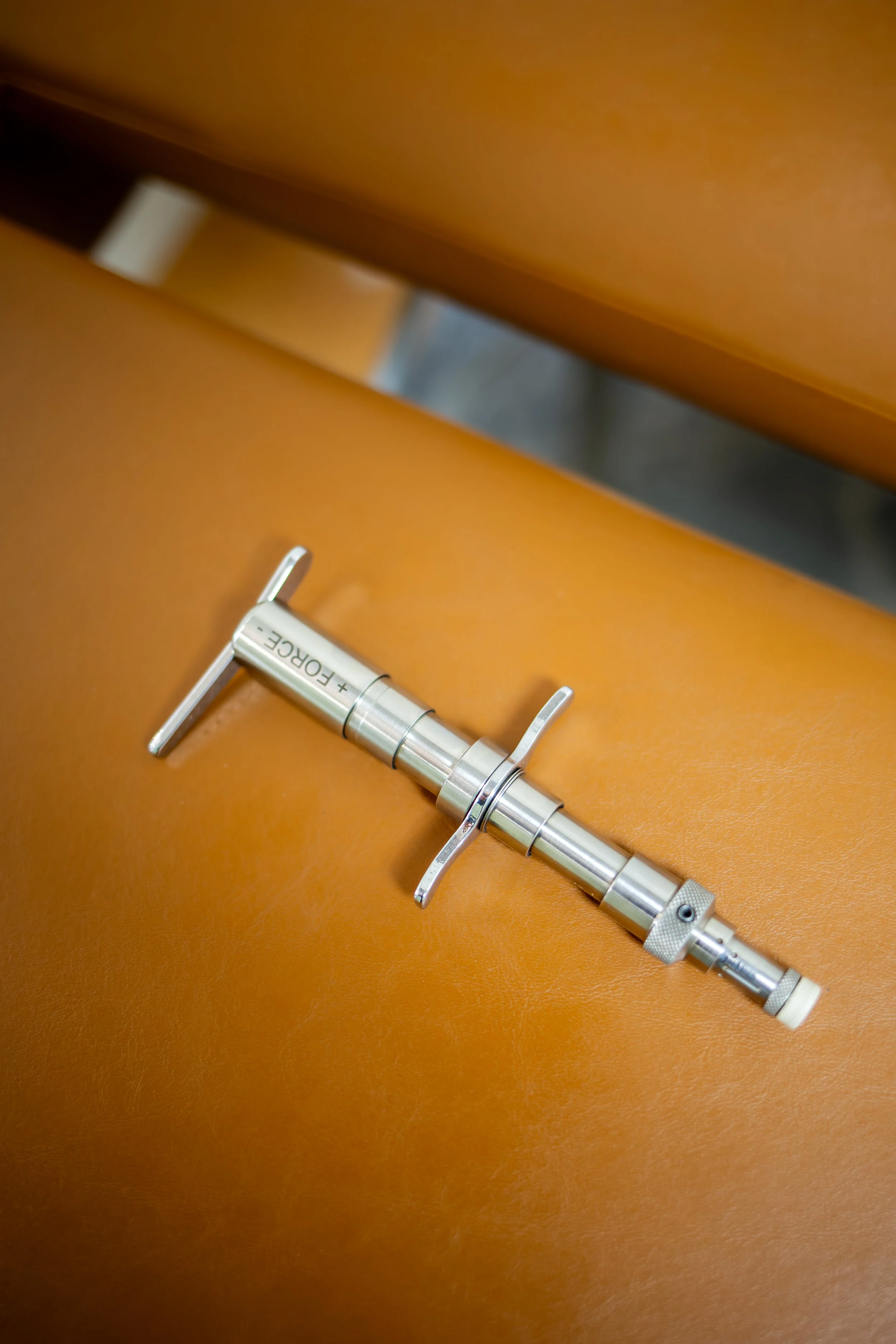 A metal syringe with the words '+' and 'FORCE' engraved on it, lying on an orange-brown leather surface.