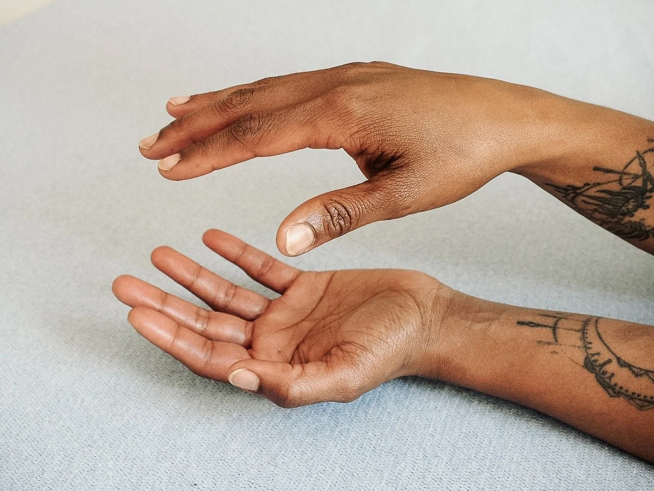 Nahaufnahme von zwei tätowierten Händen, eine schwebt über der anderen, auf einem hellen Stoffhintergrund.