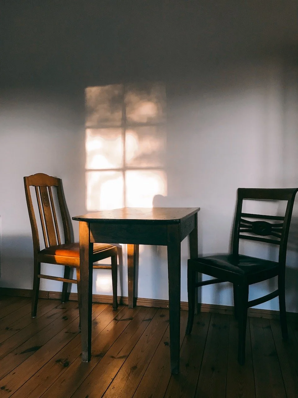 Ein Holztisch mit zwei Stühlen vor einer weißen Wand, durch die Sonnenlicht und Schatten eines Fensters fällt.