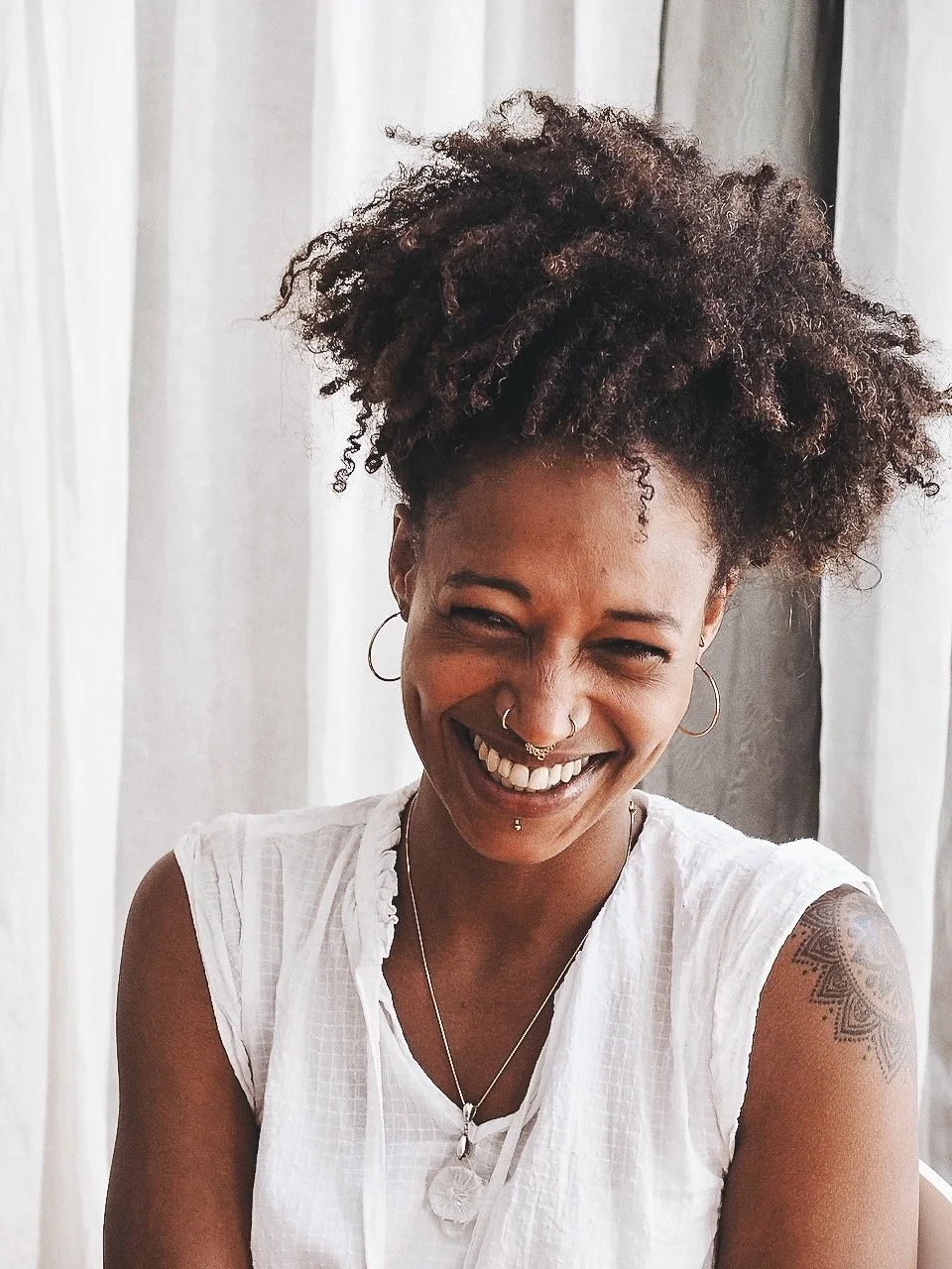 Lächelnde Frau mit dunklen, lockigen Haaren, Piercings im Gesicht, Tattoos am Arm und Schmuck, trägt ein weißes Oberteil vor einem Hintergrund aus weißen Vorhängen.