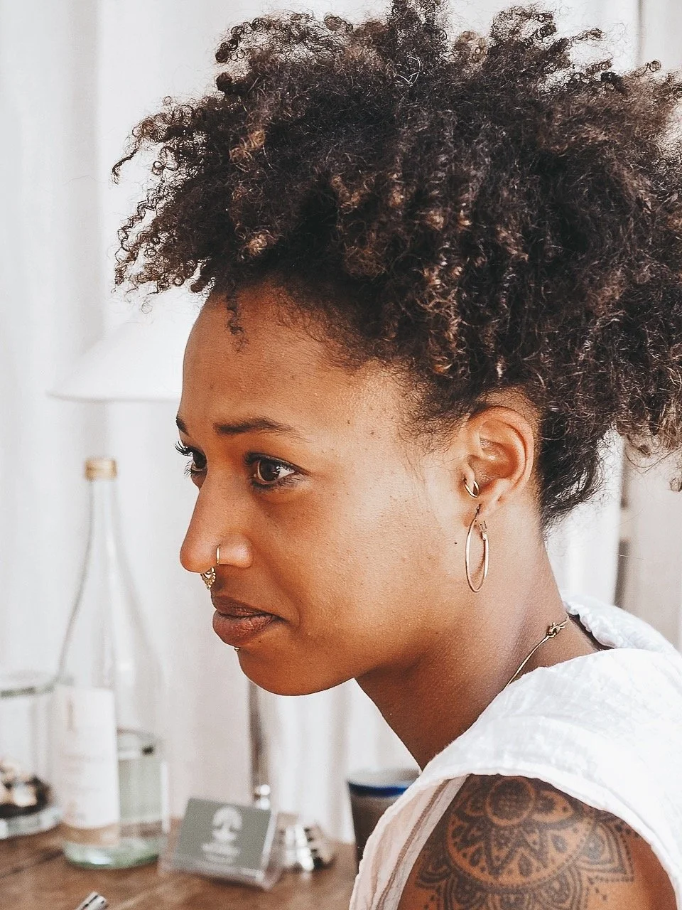 Junge Frau mit dunklen, lockigen Haaren und Piercings, trägt Ohrringe und ein weißes Top, im Hintergrund Lampen und Wasserflaschen.