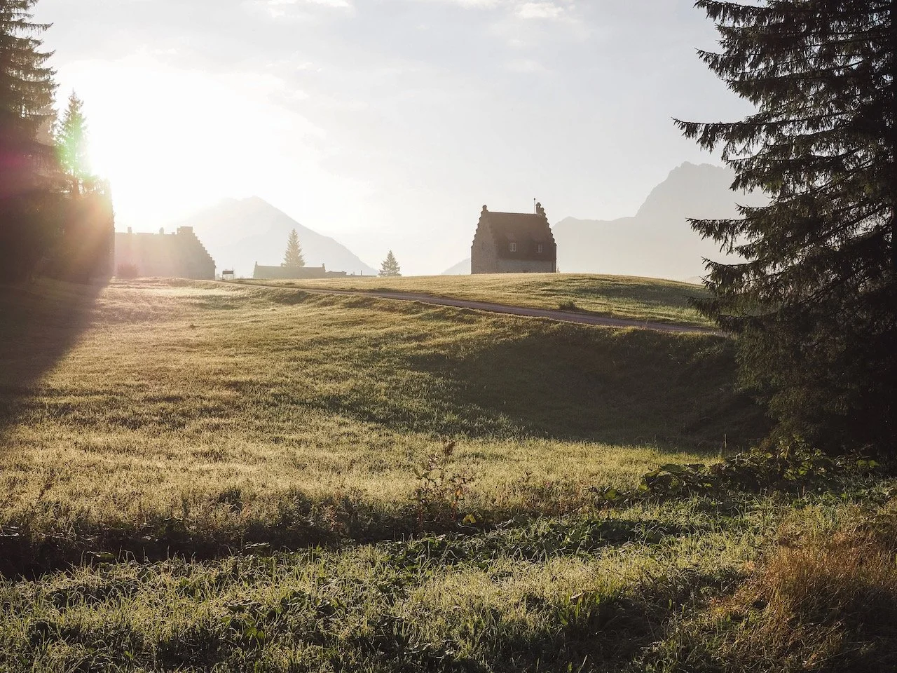 Landschaft mit einem Haus auf einer Anhöhe, umgeben von Bäumen und Bergen im Hintergrund, sonniger Himmel