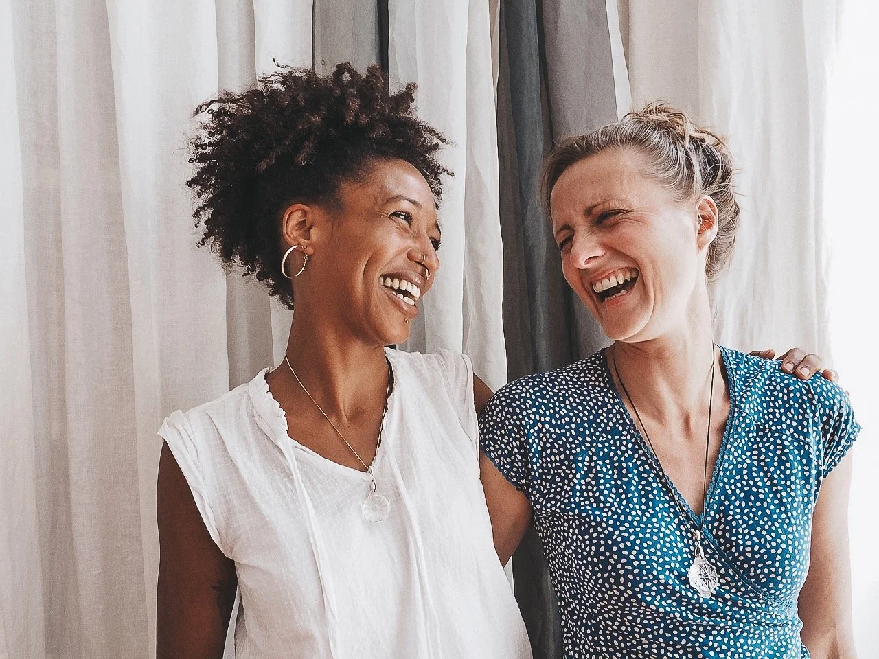 Zwei lachende Frauen, die sich umarmen, vor einem Vorhang.