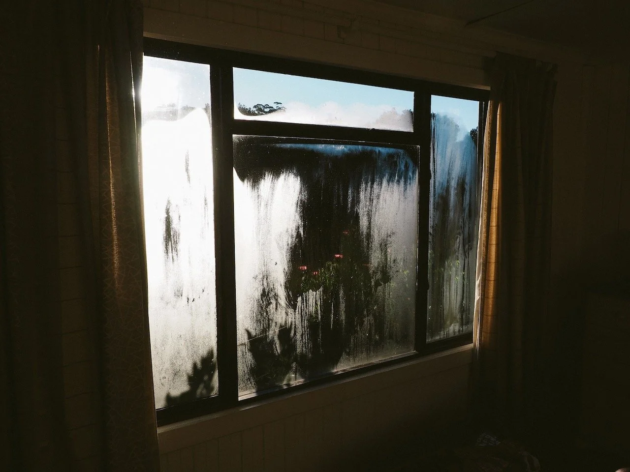 Beschlagenes Fenster, Blick nach draußen auf einen blauen Himmel und Bäume, Vorhänge an den Seiten.