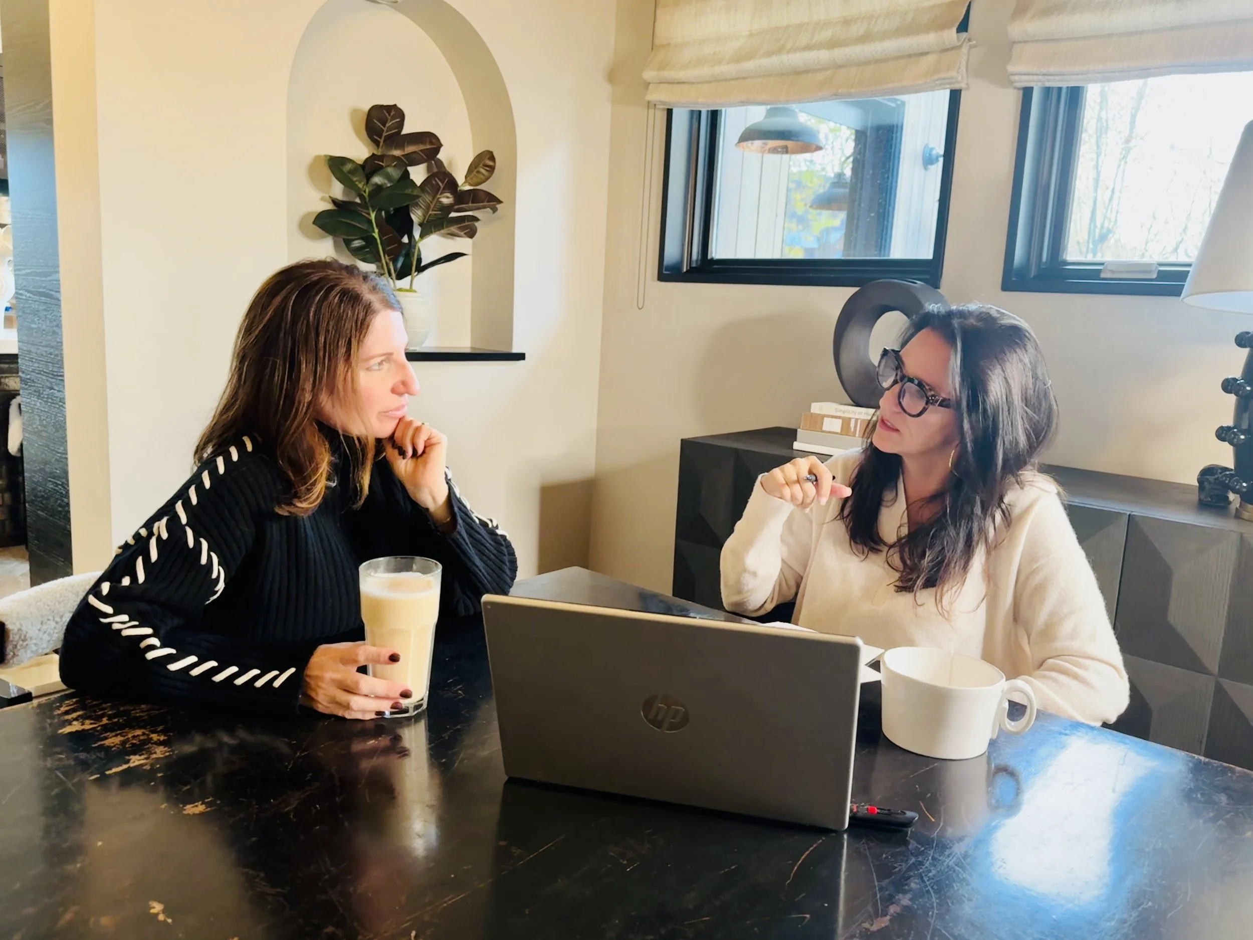 Two women having a conversation at a table with coffee mugs, a laptop, and a glass of beverage in a cozy room with natural light, a potted plant, and decorative items.
