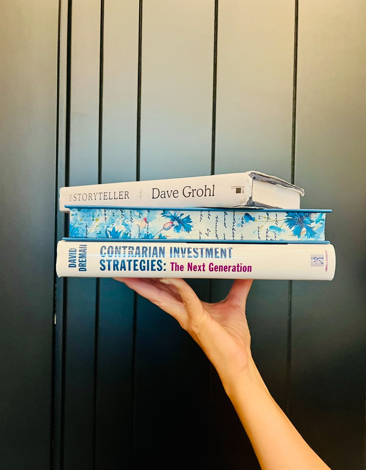 Hand holding three books stacked against a black wall. The titles are 'The Storyteller' by Dave Grohl, an unidentified patterned book, and 'Contrarian Investment Strategies: The Next Generation' by David Dreman.