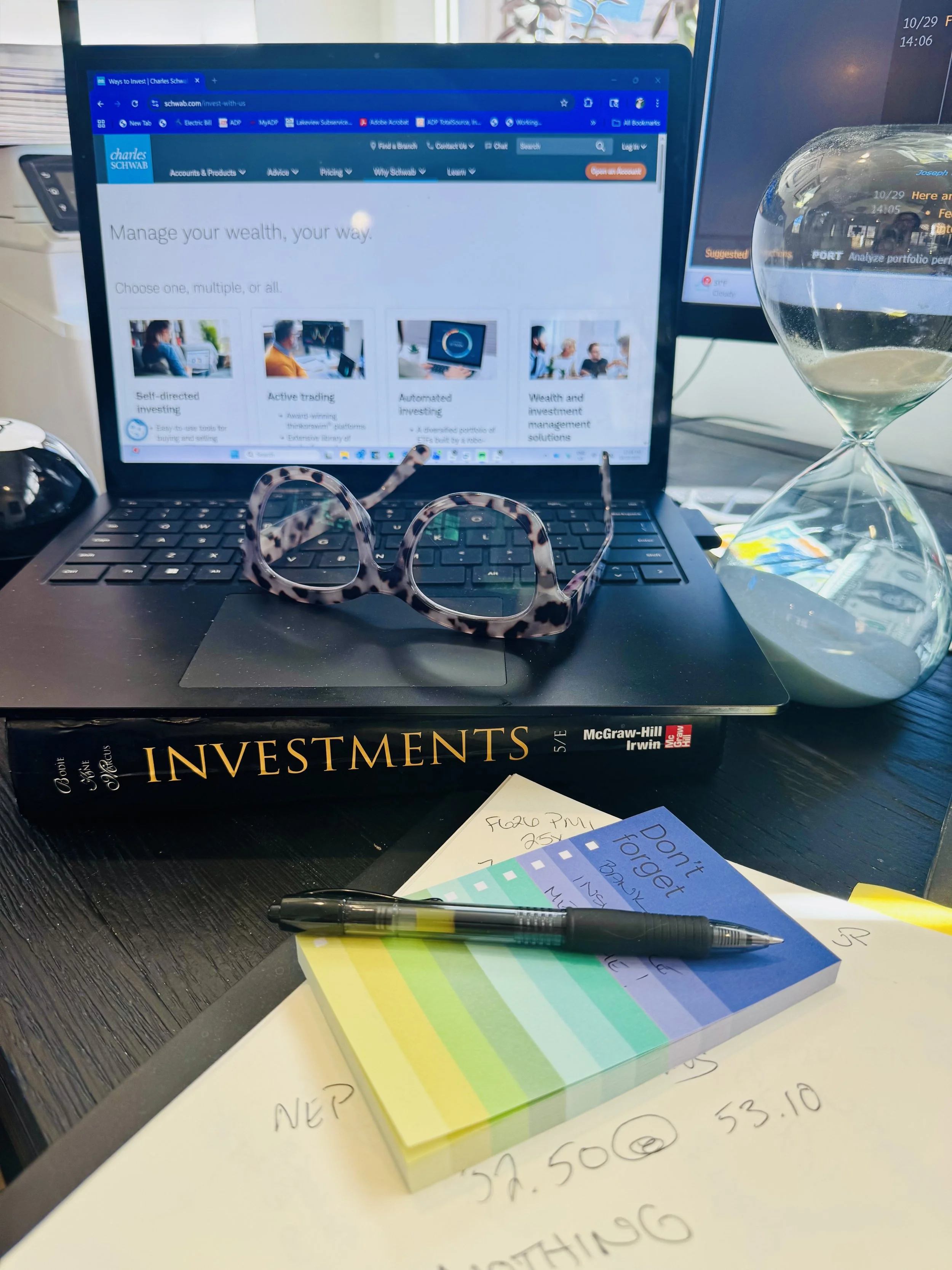 A workspace featuring an open laptop displaying a financial website, a pair of tortoiseshell glasses resting on an investment book titled 'Investments' by McGraw-Hill, a black pen on colorful sticky notes, and handwritten notes on paper, with an hourglass and a pair of sunglasses on a black wooden desk.