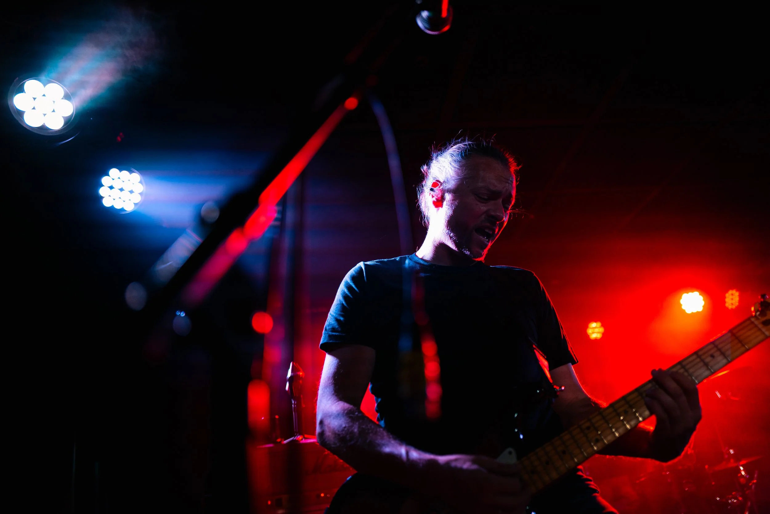 Stefan von Endzeitparty spielt Gitarre auf einer Bühne bei dunklem Licht, umgeben von bunten Scheinwerfern, die rote, blaue und weiße Lichter werfen.