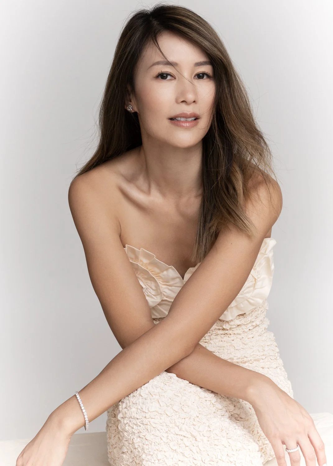 A woman with long brown hair, wearing a beige strapless dress with ruffles, sitting against a plain white background.