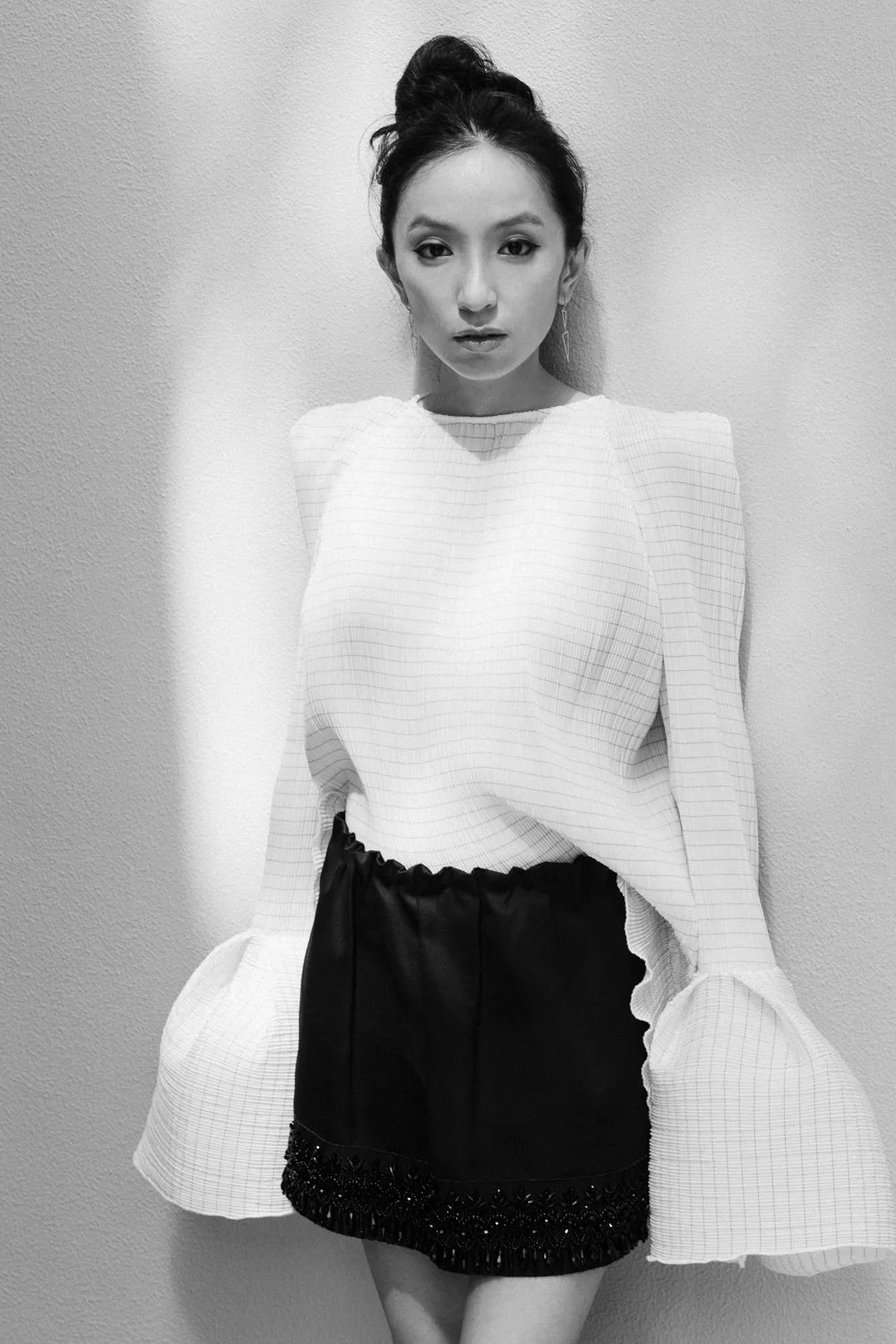 Black and white photo of a woman with her hair in a bun, wearing a white blouse with puffy sleeves and a black skirt, standing against a wall.