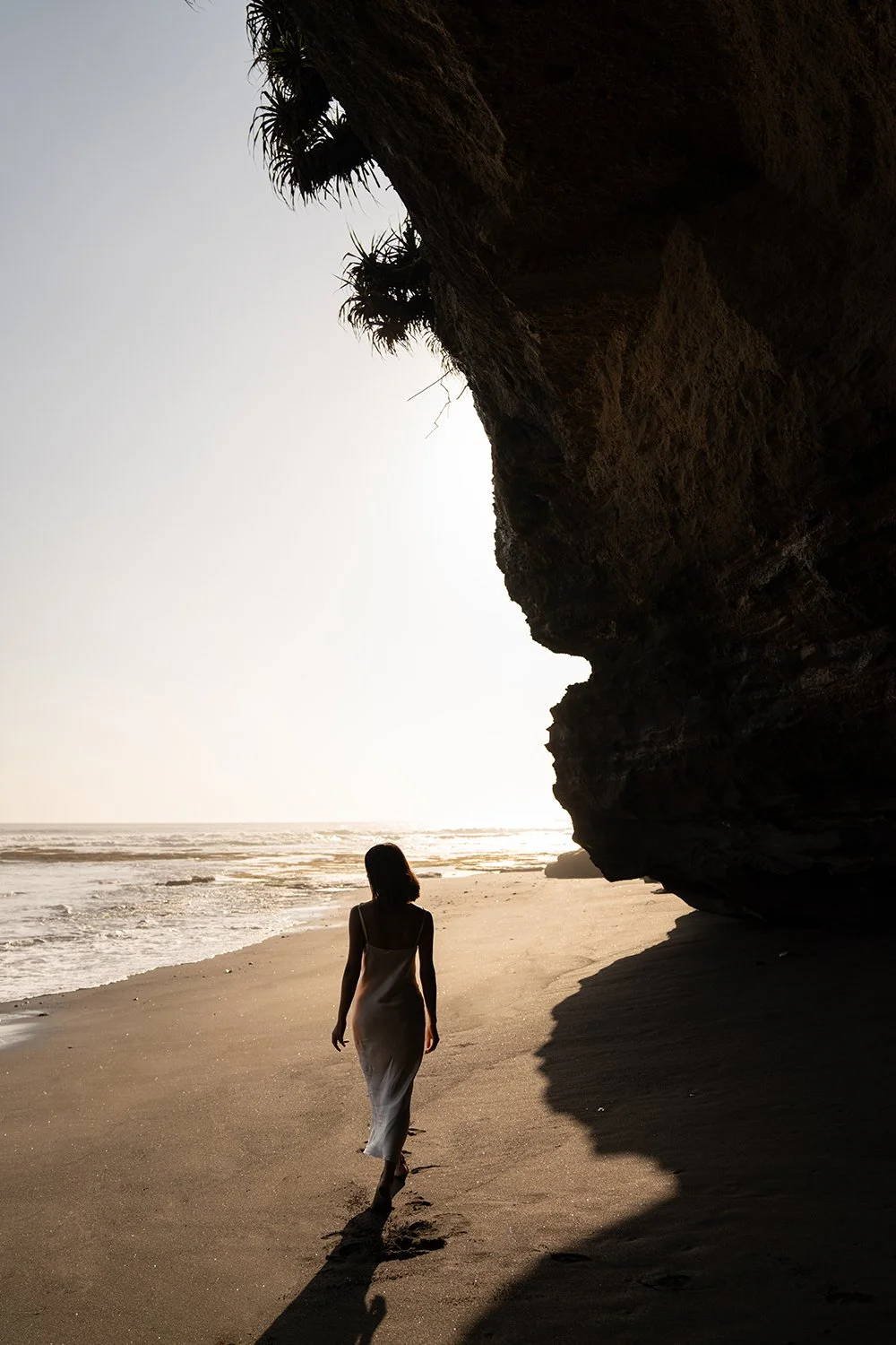 A woman walks along a sandy beach near a large rocky cliff during sunset or sunrise, casting a long shadow.