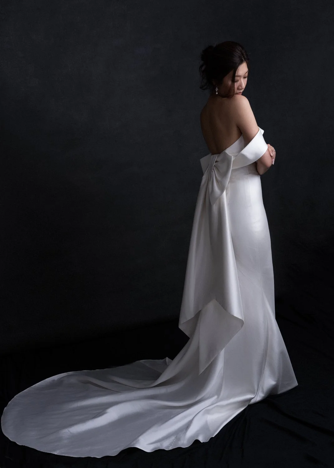A woman in a white strapless wedding dress with a long train stands against a black background, looking down with her arms crossed.