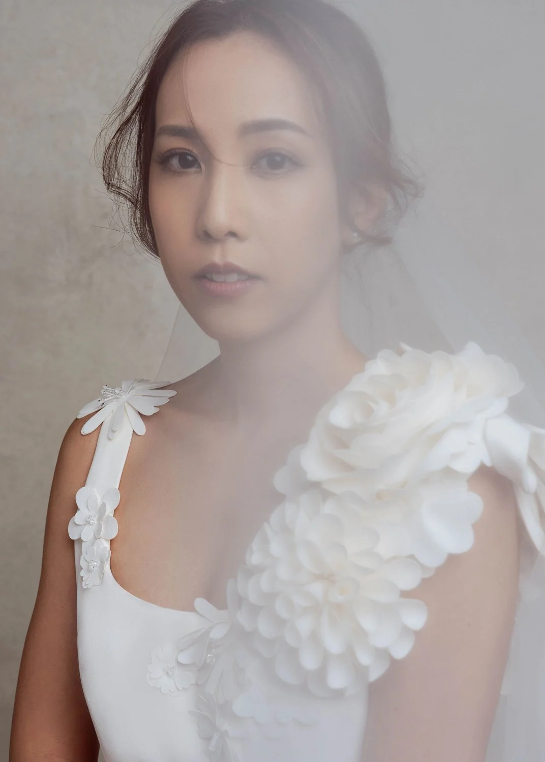 A woman with short wavy brown hair wearing a white dress with large floral embellishments on the shoulder.