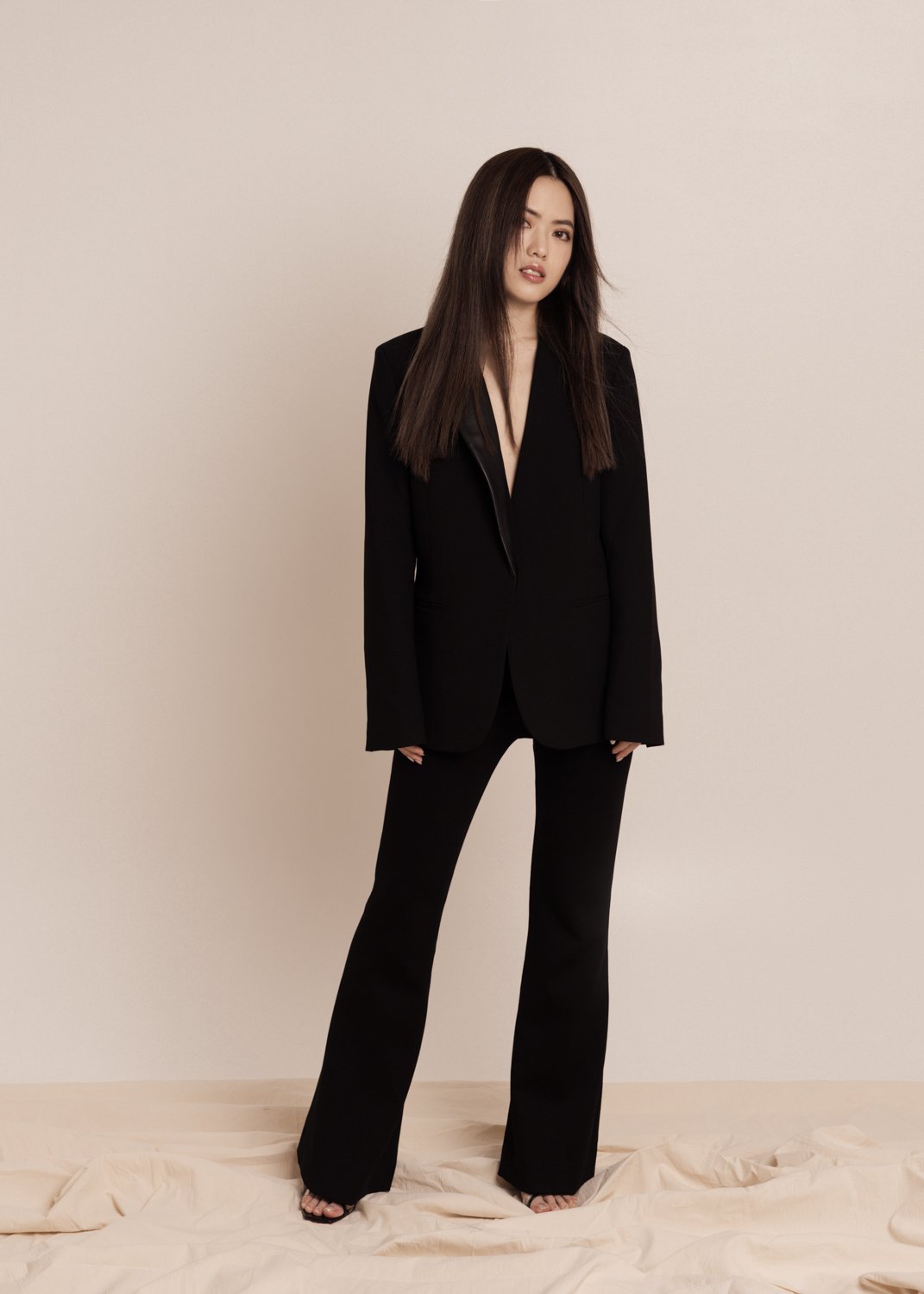 A woman in a black suit standing against a plain beige background.