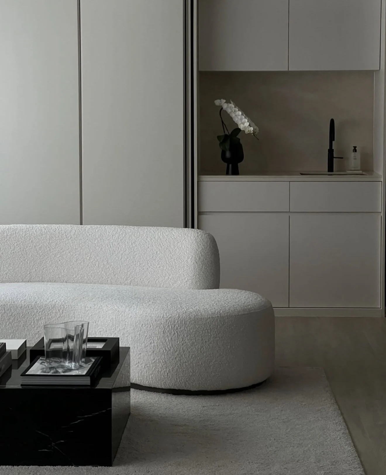 Modern living room with white textured sofa, black coffee table, clear glass, and books, with a minimalist kitchen area in the background featuring a black faucet, white cabinetry, and a flower vase with white orchids.
